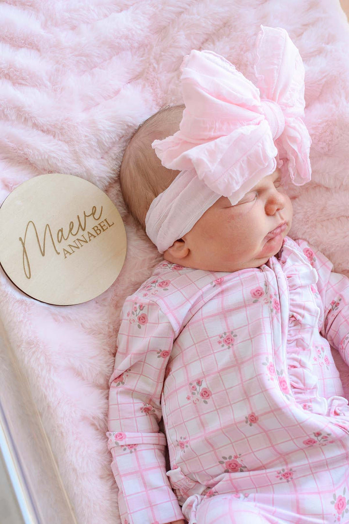 Sleeping newborn girl wearing a Caden Lane pink floral windowpane sleeper gown and pink bow headband, lying on pink faux fur next to a Caden Lane engraved wood sign reading "Maeve Annabel."