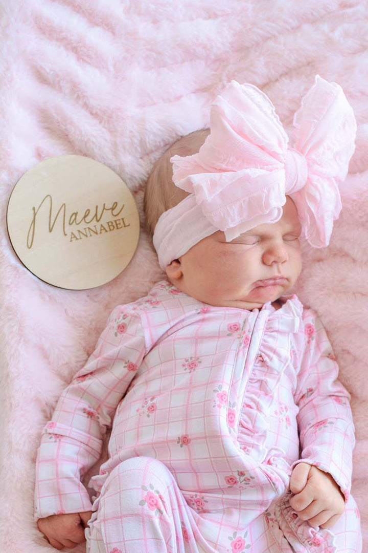 Overhead view of a newborn in a Caden Lane pink floral windowpane footie gown with ruffle trim, sleeping beside a wooden "Maeve Annabel" name sign in a hospital bassinet.