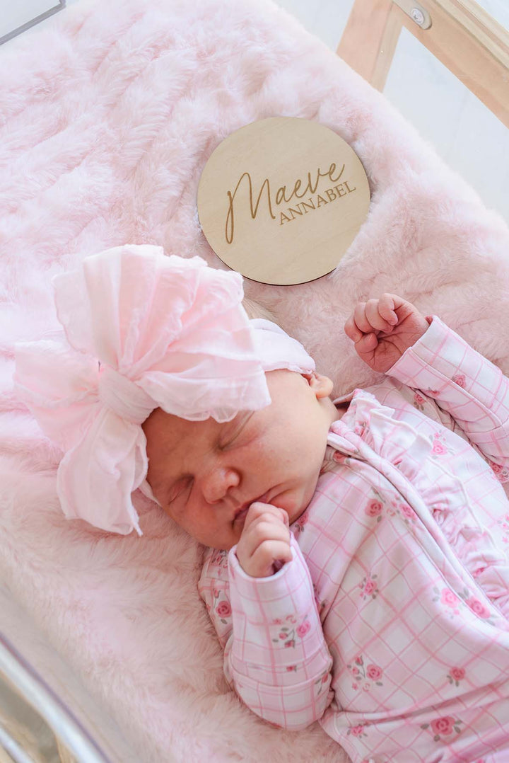 Close-up of a sleeping newborn in a Caden Lane pink bow headband and floral windowpane sleeper gown, positioned beneath a wooden "Maeve Annabel" birth announcement sign on pink faux fur.
