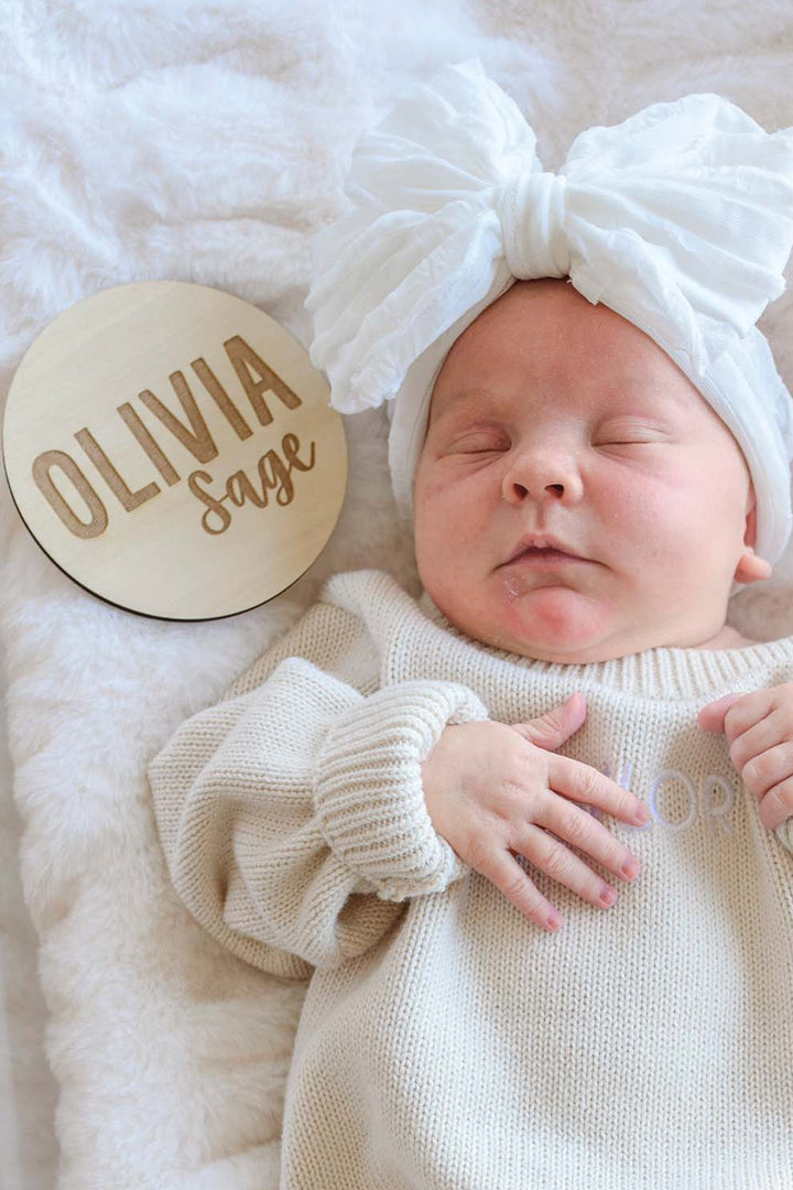 Close-up of a sleeping newborn in a Caden Lane white bow headband and cream knit romper, positioned next to a wooden "Olivia Sage" birth announcement sign on a fuzzy cream blanket.