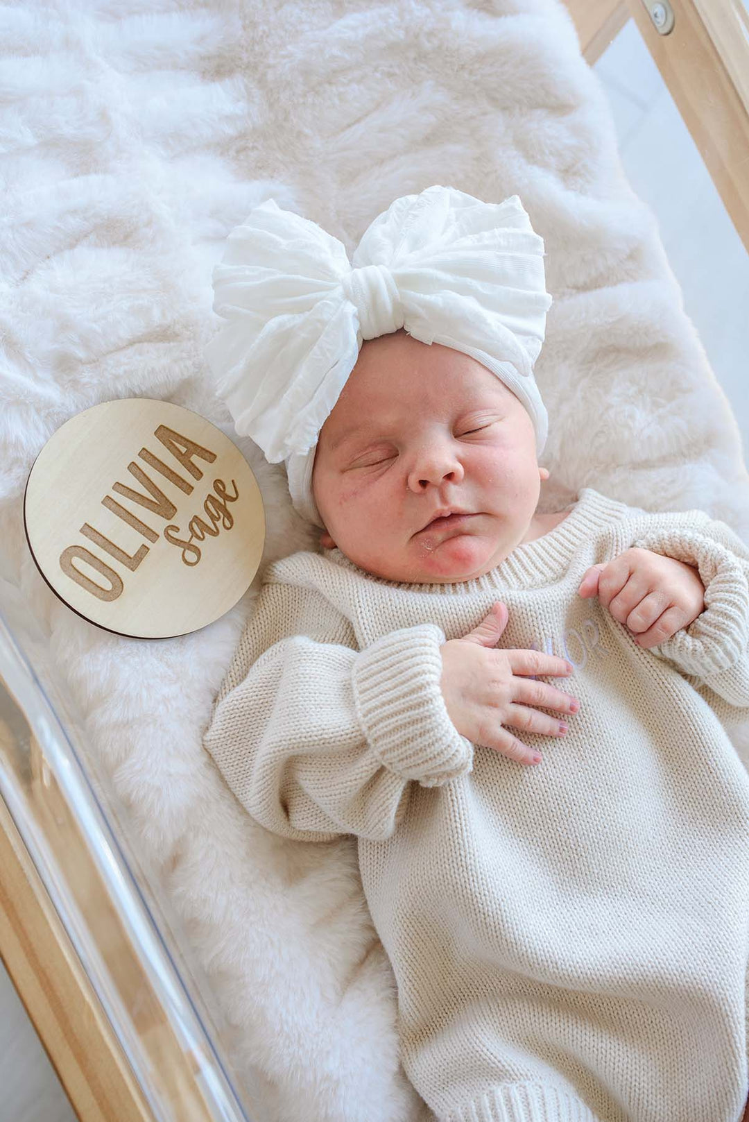 Sleeping newborn wearing a Caden Lane white bow headband and cream knit romper, lying in a hospital bassinet next to a Caden Lane engraved wood sign reading "Olivia Sage."