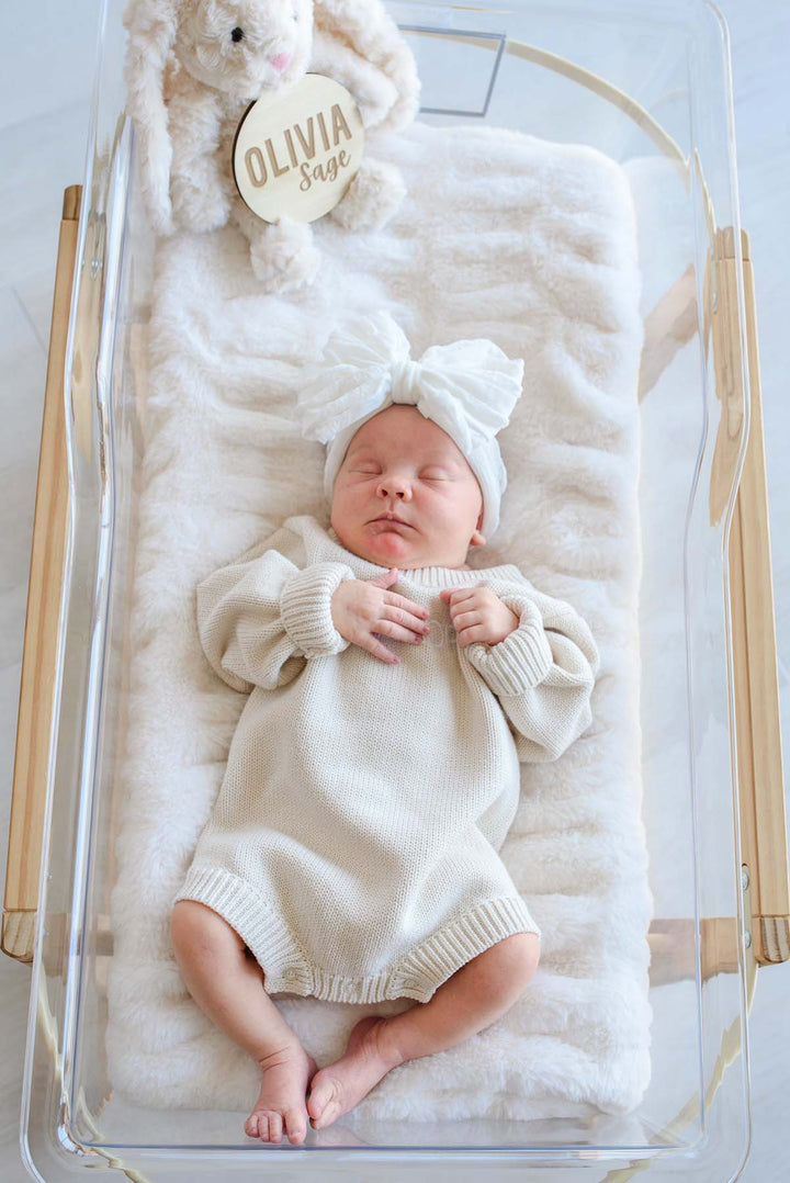 Newborn girl in a Caden Lane white bow headband and cream knit outfit resting in a clear hospital bassinet, with a wooden "Olivia Sage" name sign and plush bunny beside her.