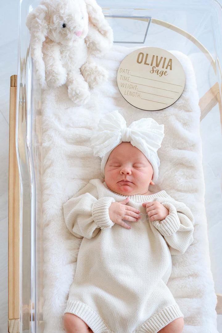 Overhead view of a newborn in a cream knit outfit and Caden Lane white bow headband, resting beside a personalized wooden "Olivia Sage" sign and plush bunny in a hospital bassinet.