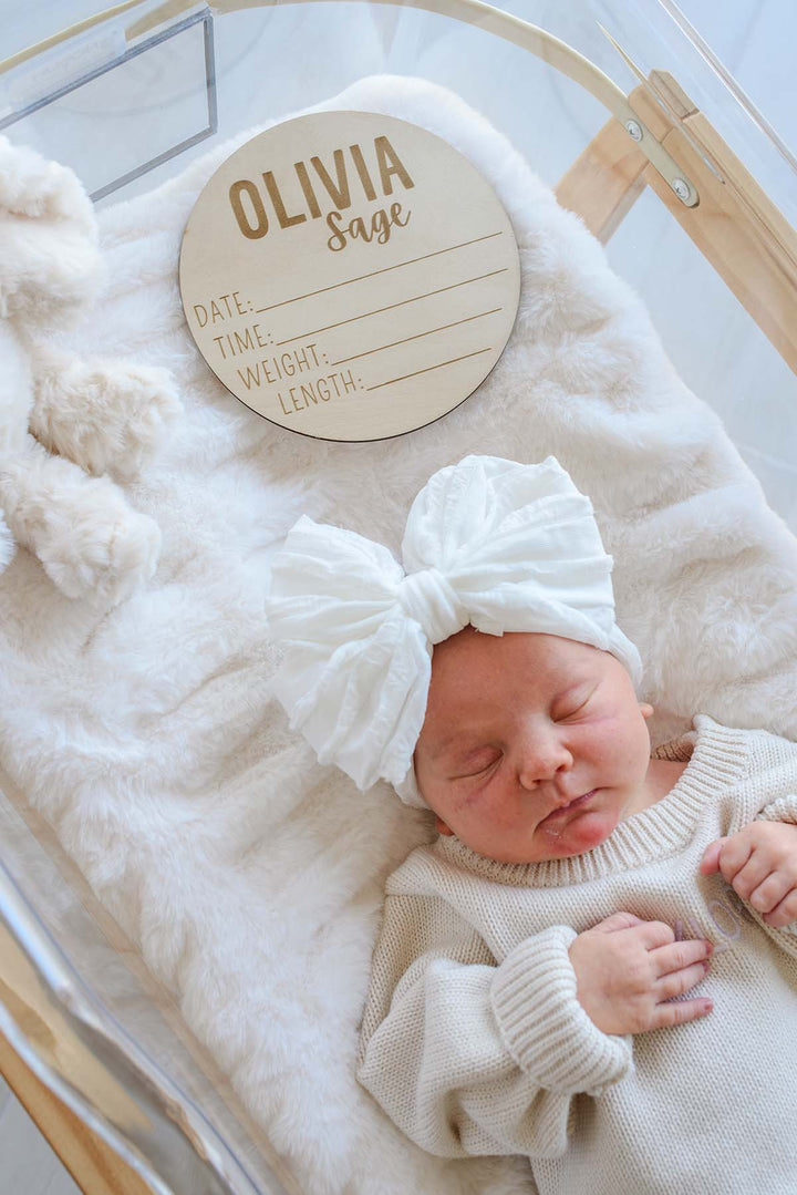 Sleeping newborn in a cream knit sweater and white bow headband, lying in a hospital bassinet beside a Caden Lane personalized wooden birth announcement sign reading "Olivia Sage" with blank date, time, weight, and length lines, plus a plush bunny.
