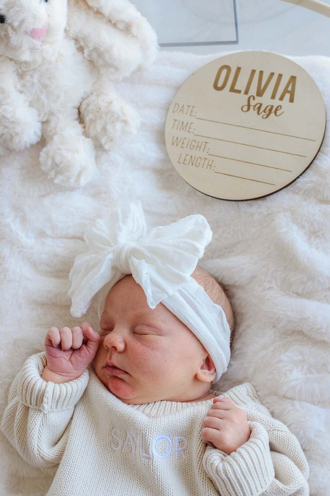 Newborn wearing a Caden Lane white ruffle bow headband and cream knit "Saylor" sweater, sleeping next to a Caden Lane engraved wood name sign reading "Olivia Sage" and a cream bunny stuffed animal.