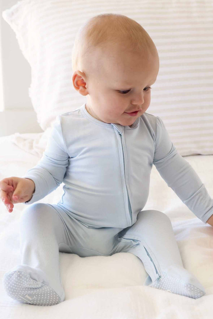 A baby sitting on a white bed wearing a light blue long-sleeve zip-front sleeper with foot covers.