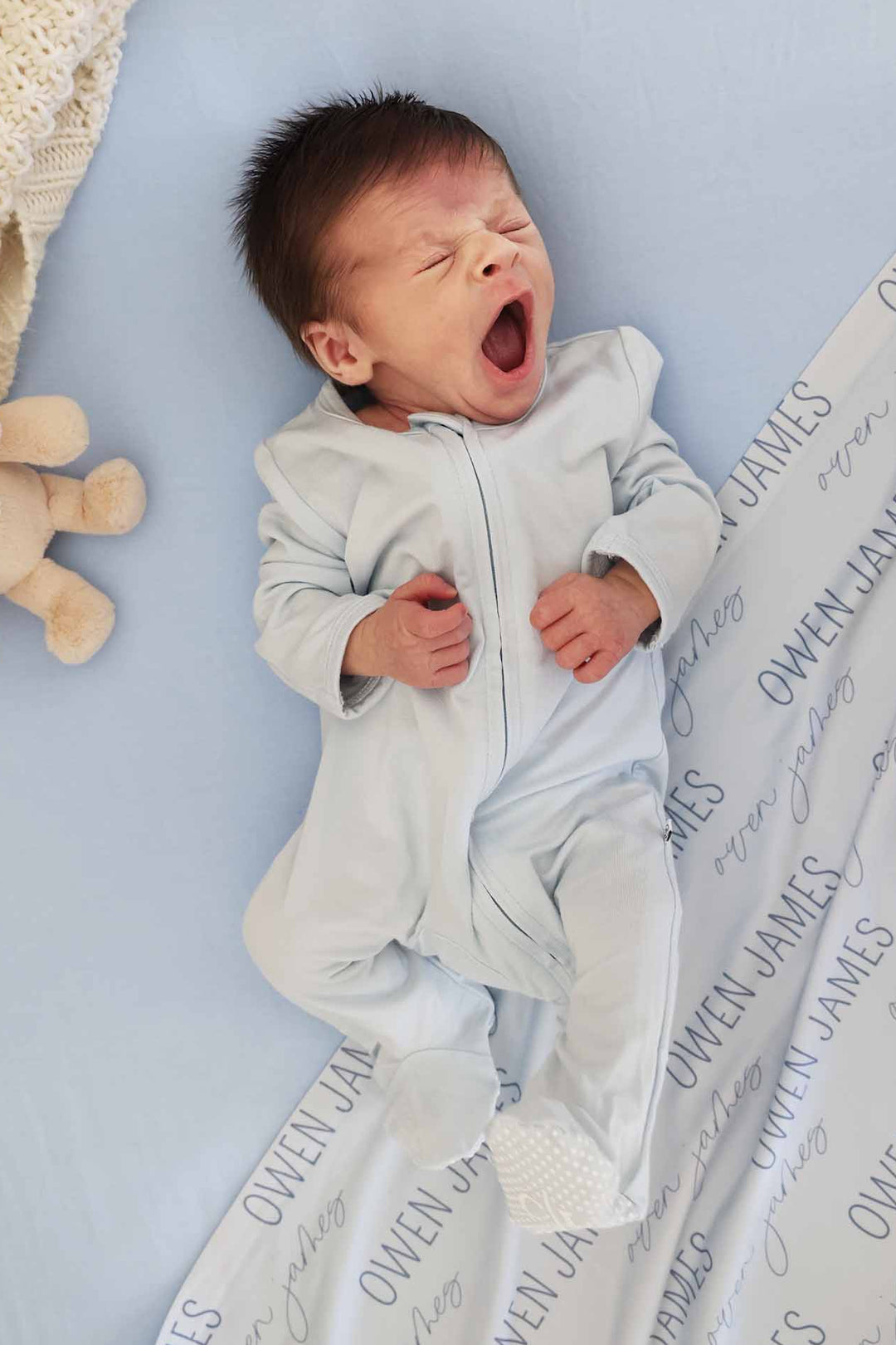 yawning baby boy in light blue zipper footie pajama with matching personalized swaddle in background 
