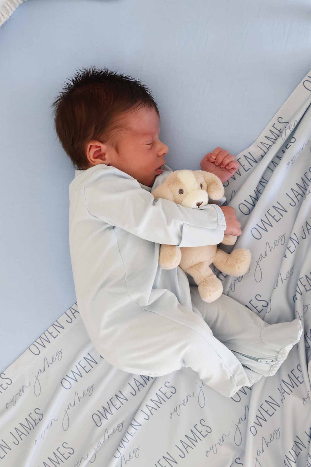 baby boy laying on his side holding a dog stuffed animal laying on a blue crib sheet and matching personalized swadle blanket 