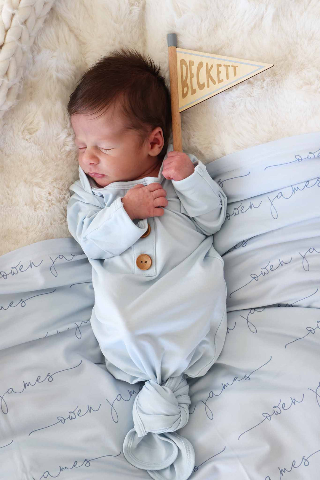 baby boy laying down in light blue newborn gown holding a flag name sign 