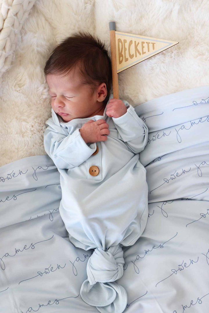 baby boy laying down in light blue newborn gown holding a flag name sign 