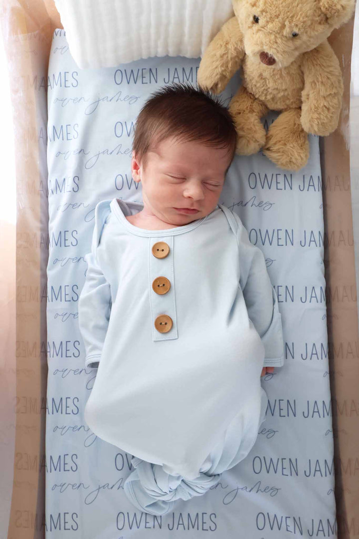 baby boy wearing a light blue long sleeve newborn knot gown on top of  matching personalized swaddle blanket with a teddy bear in bassinet 