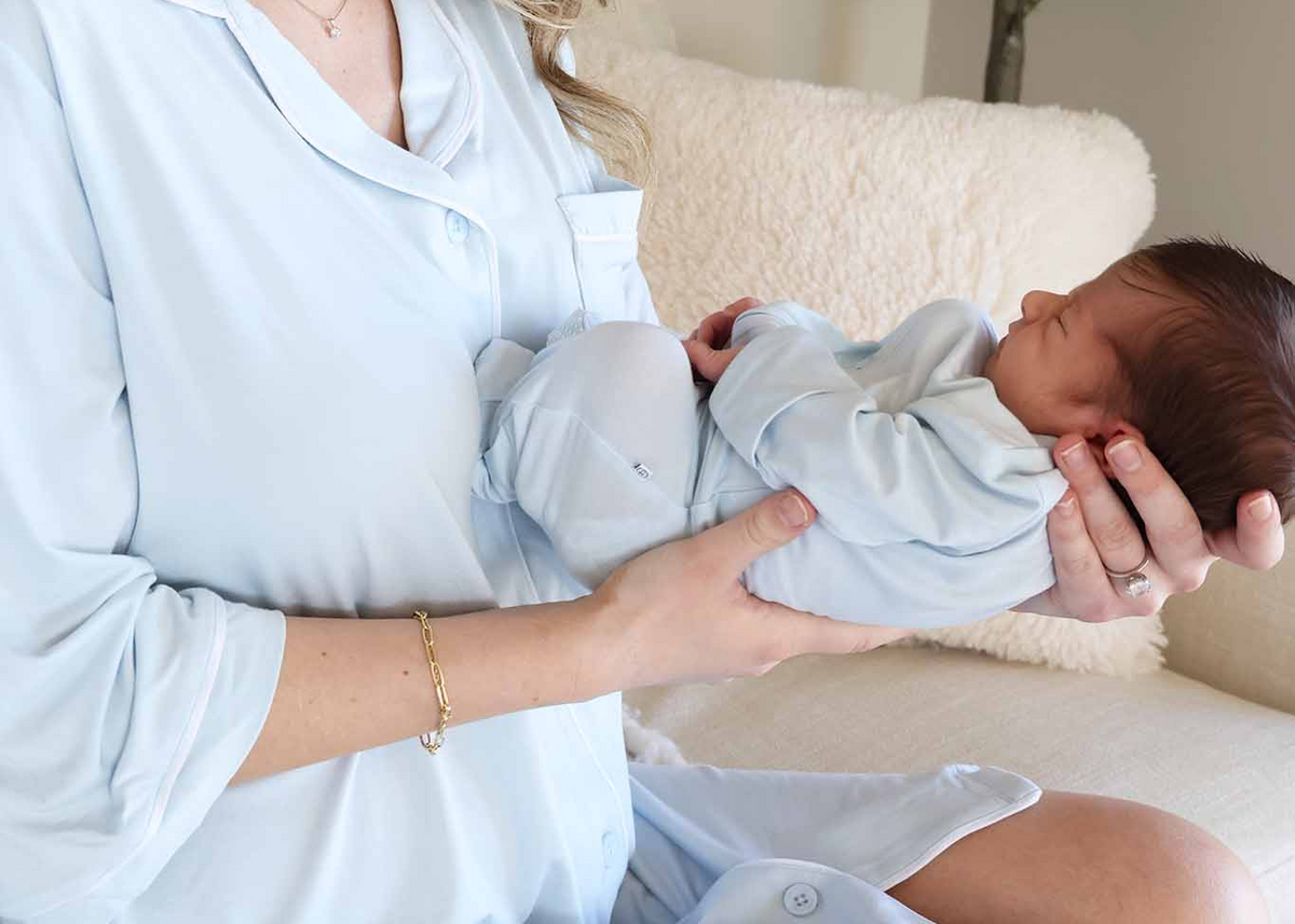 Cropped shot of a mother cradling her sleeping newborn on a sofa, wearing a Caden Lane baby blue labor and delivery nightgown, paired with the baby's matching blue outfit.