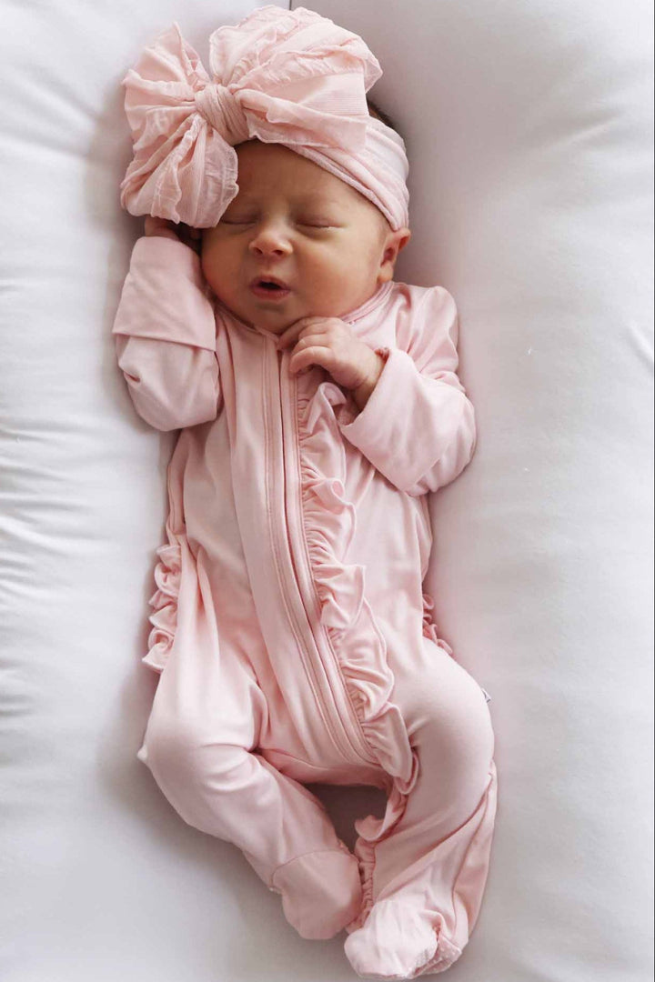 Newborn baby in a pink outfit with a matching headband lying on a white pillow.