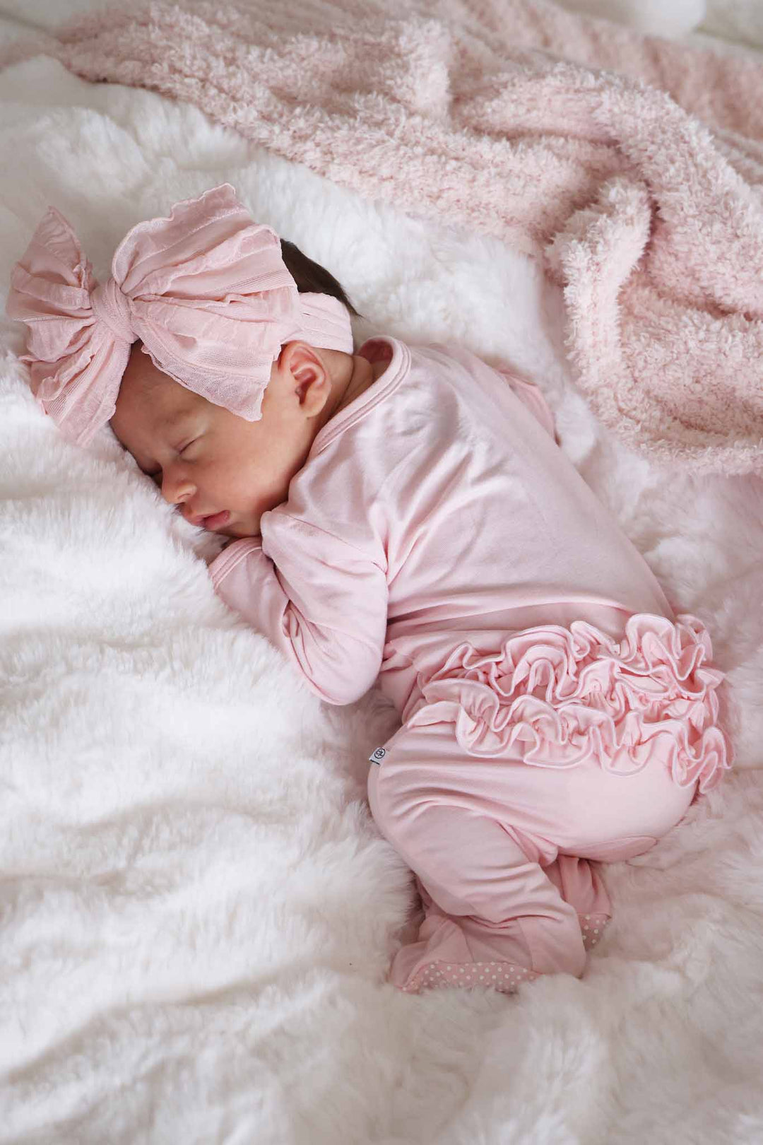 baby girl laying on stomach wearing the baby pink ruffle buttom zipper footie with matching large ruffle bow 