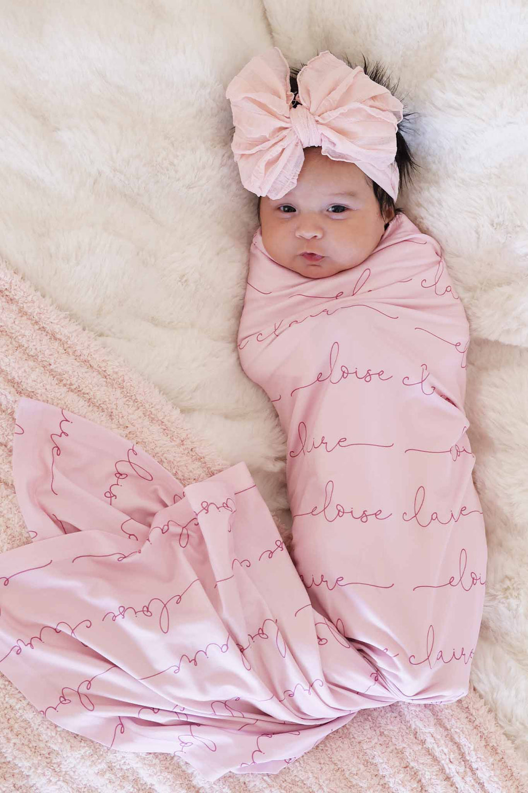 baby girl wrapped in light pink personalized swaddle blanket with dark pink font and matching ruffle bow 
