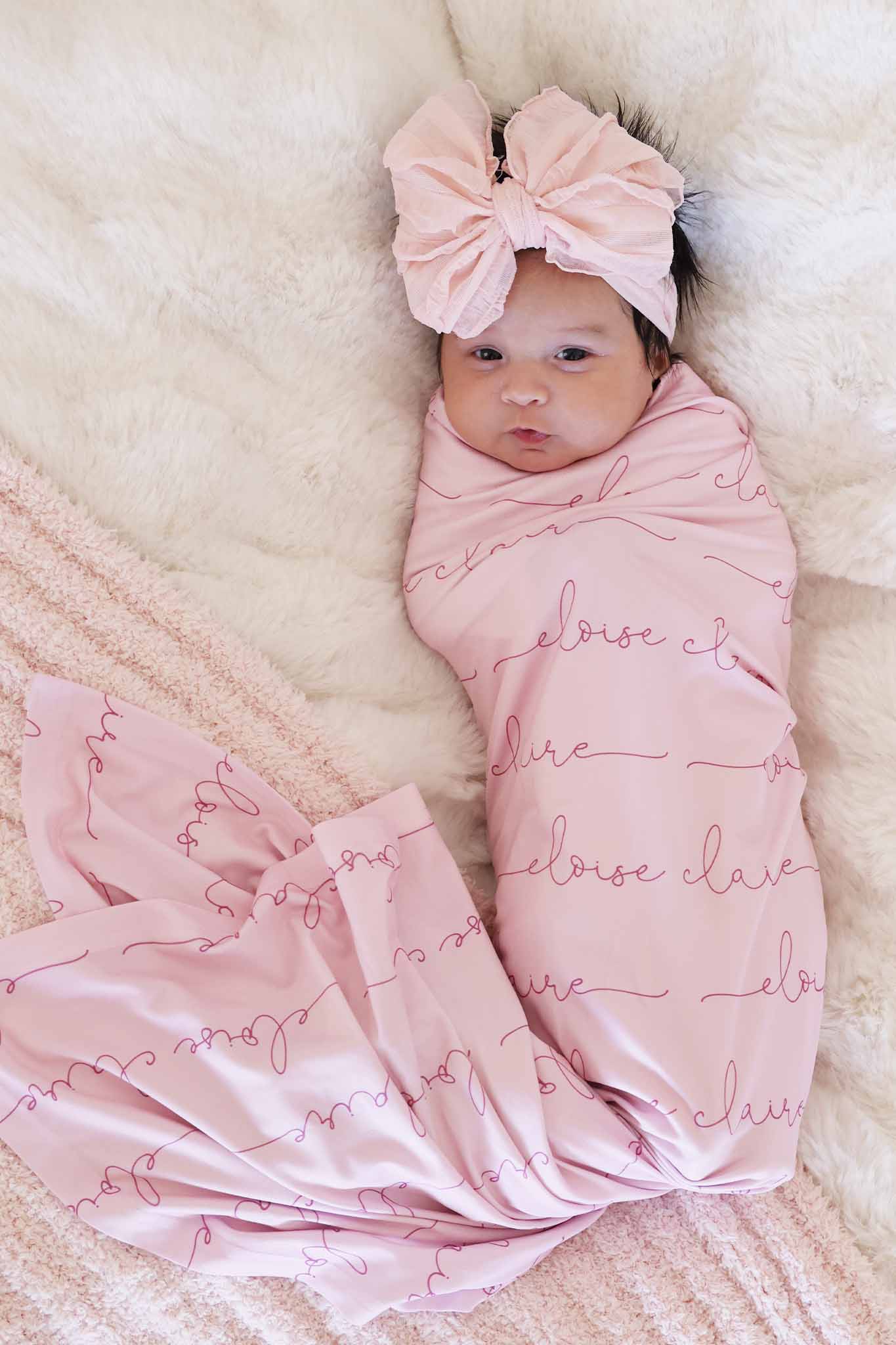 baby girl wrapped in light pink personalized swaddle blanket with dark pink font and matching ruffle bow 