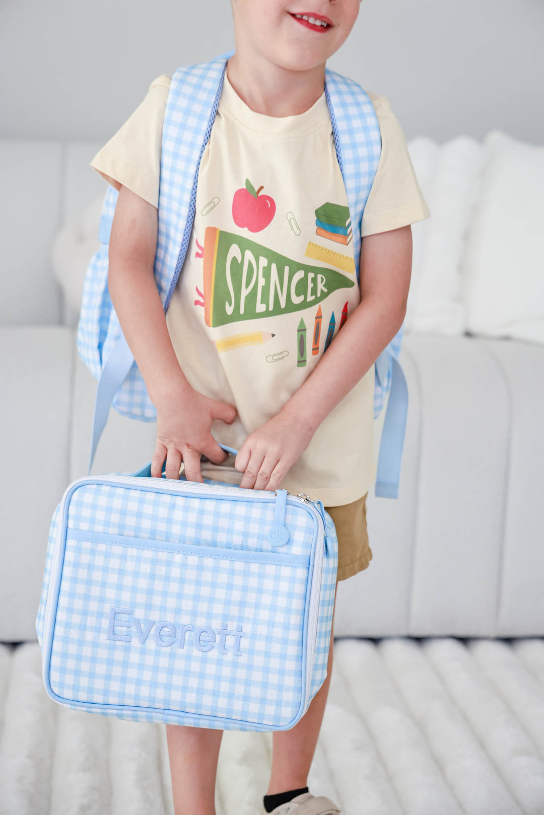 boy wearing personalized back to school tee holding a blue gingham embroidered lunchbox and wearing matching backpack