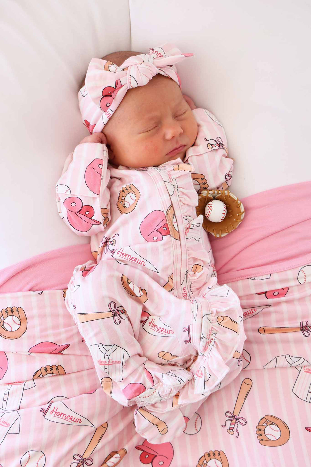 baby girl laying in lounger wearing pink ballpark babes footie with a matching headwrap and small baseball glove prop 