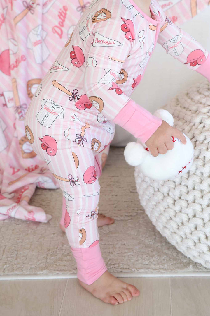 standing baby girl leaning against ottoman wearing a pink ballpark babes convertible zip romper and holding a stuffed baseball 