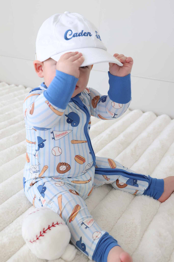 baby boy in baseball themed zip romper pajama wearing an embroidered caden lane baseball hat 