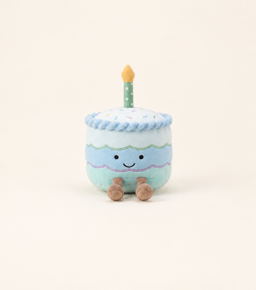 Caden Lane plush birthday cake toy with three tiers in blue, white, and mint green frosting, a smiling embroidered face, brown stubby feet, and a green polka-dot candle with an orange flame on top.