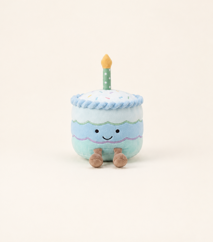 Caden Lane plush birthday cake toy with three tiers in blue, white, and mint green frosting, a smiling embroidered face, brown stubby feet, and a green polka-dot candle with an orange flame on top.