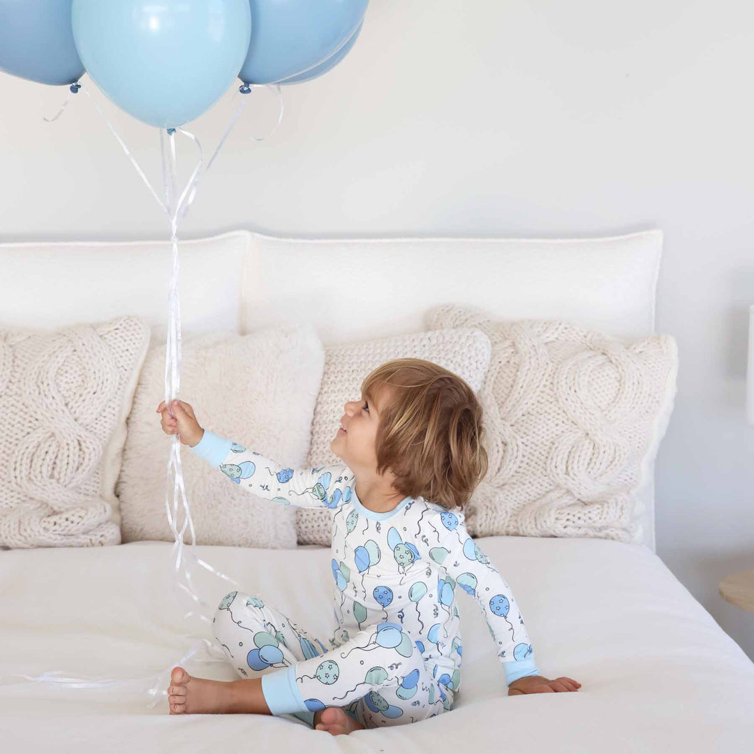 boy sitting on bed holding and looking up at balloons in blue and green balloon pajama set 