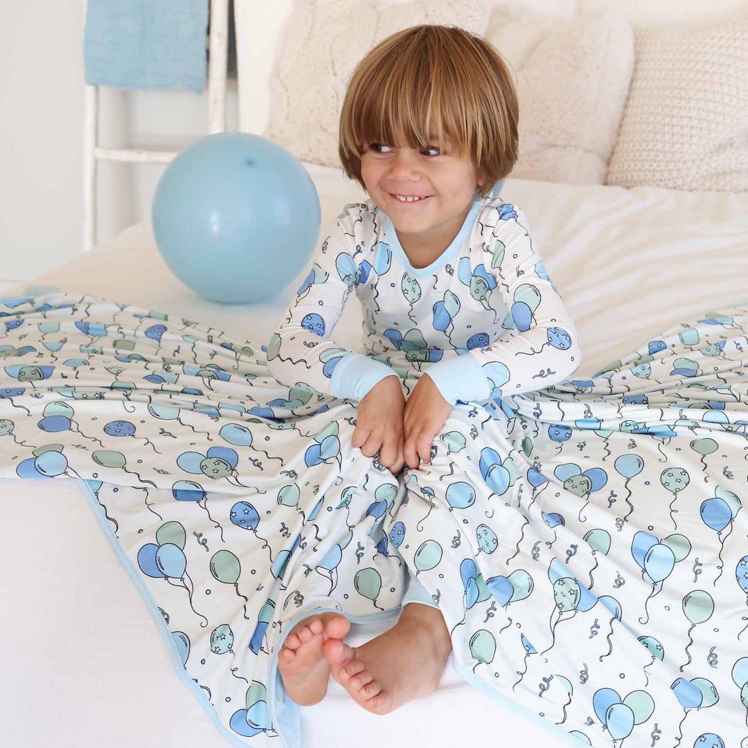 boy sitting on bed wearing blue and green balloon pajamas with a matching blanket draped over him 