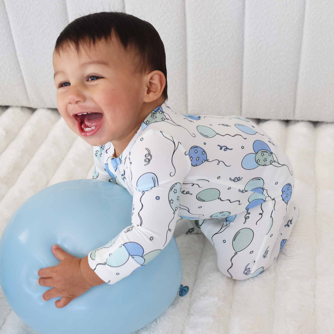 baby boy kneeling and holding balloon wearing a blue balloon footie pajama 