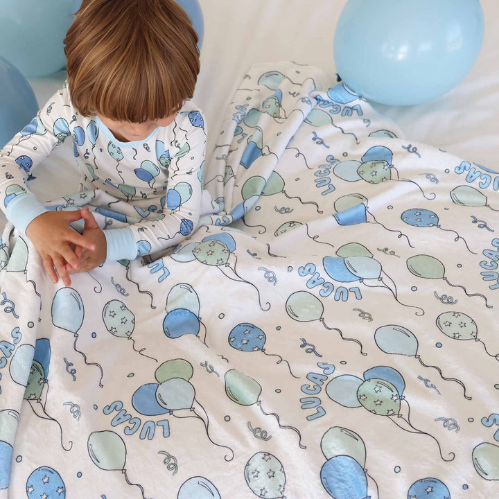 boy sitting on bed wearing  a birthday balloon long sleeve pajama set with a matching personalized blanket across his lap
