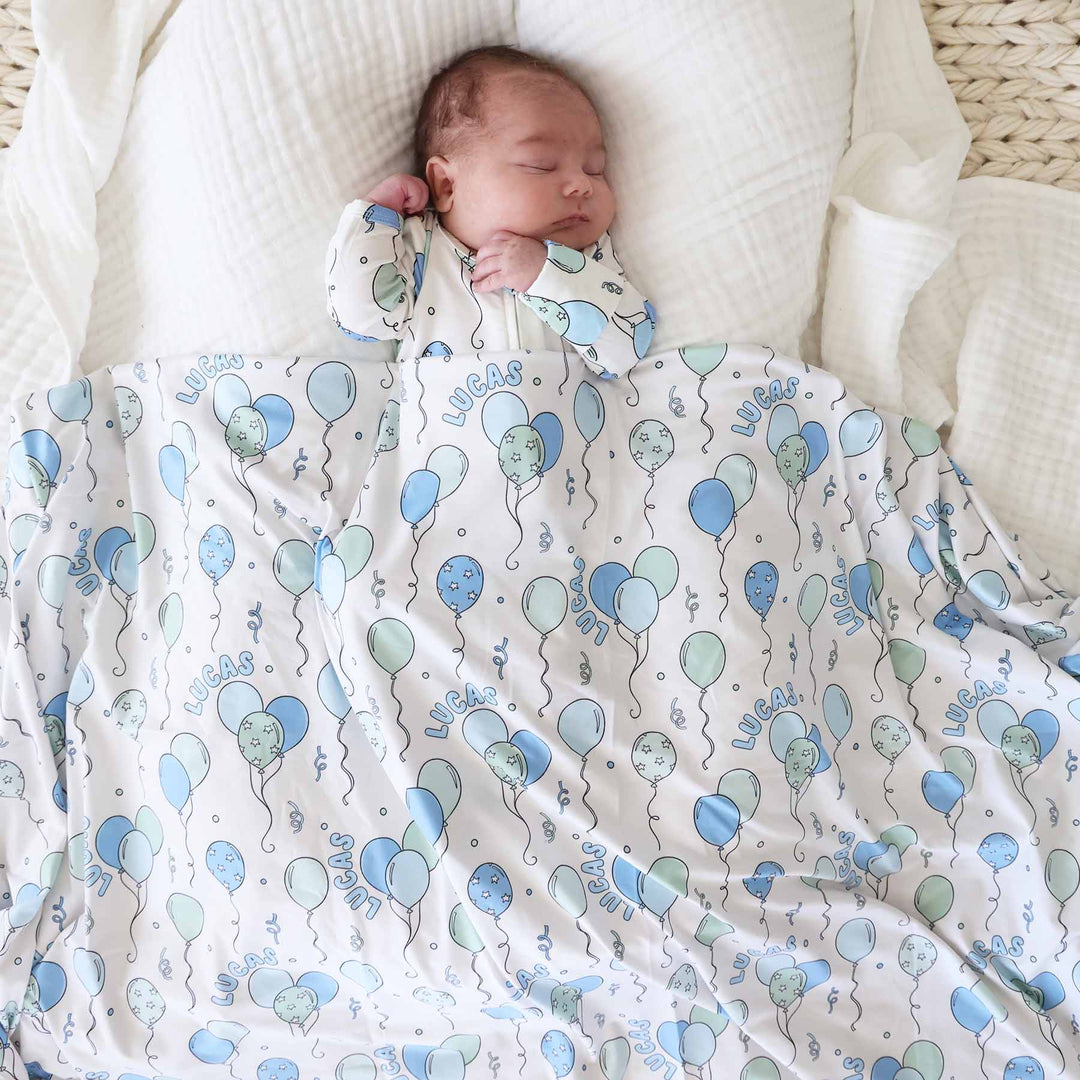 baby boy laying down in lounger with blue and green balloon footie pajama covered with matching personalized swaddle blanket 