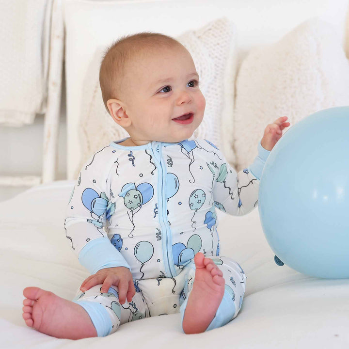 baby sitting on bed wearing blue and green balloon pajama romper next to a blue balloon 