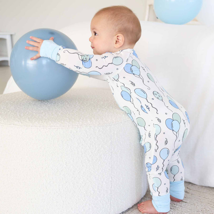 standing boy wearing balloon and streamer pajama romper reaching for a blue balloon