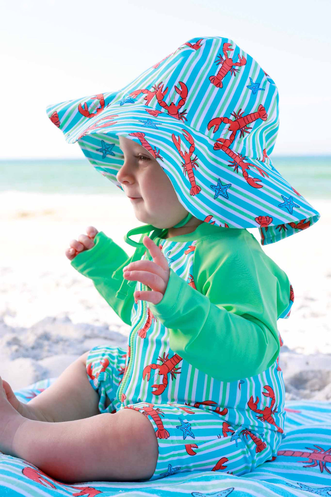 baby boy sitting on beach in lobster sun hat and matching shorty romper