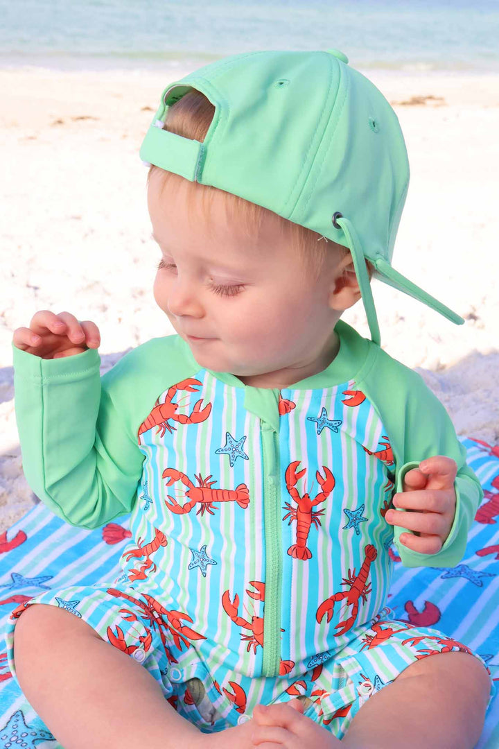 boy sitting on the beach in lobster shorty swim romper with a matching bright green surf hat 