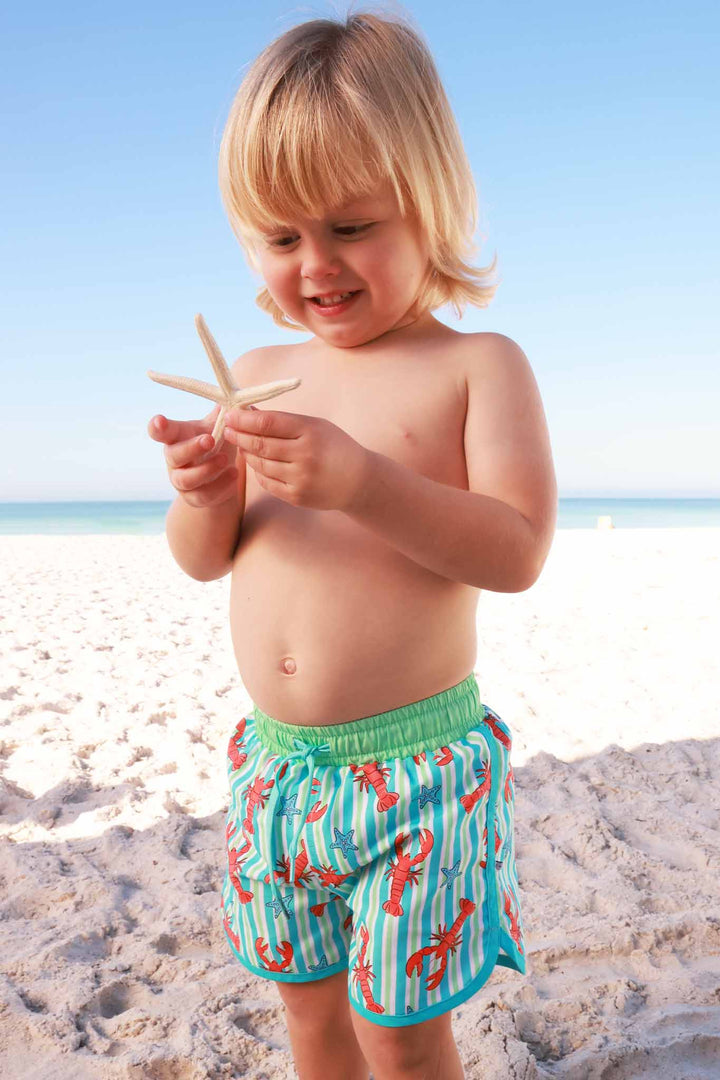 boy standing on beach playing with starfish wearing lobster swim trunks 