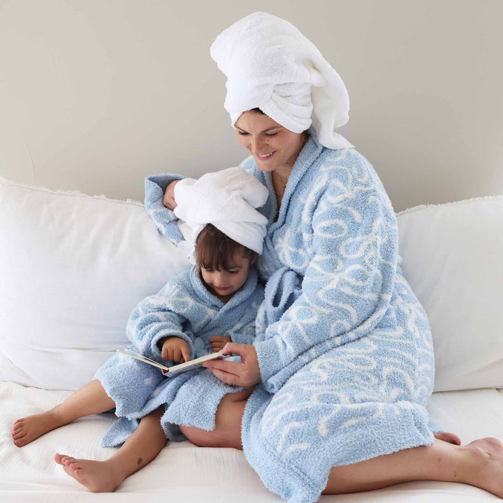 mom and daughter sitting on a couch reading a book wearing matching plush robes that are blue with white bows with towels wrapped on their heads 