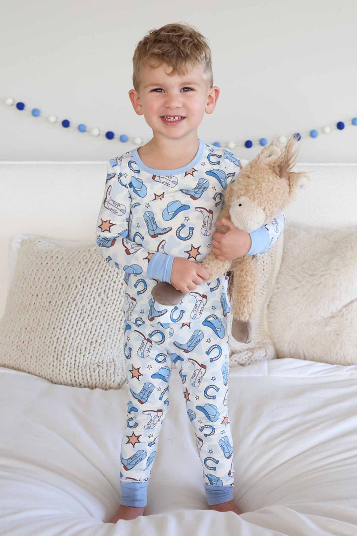 boy standing on bed in blue cowboy themed pajama set holding a horse stuffed animal 