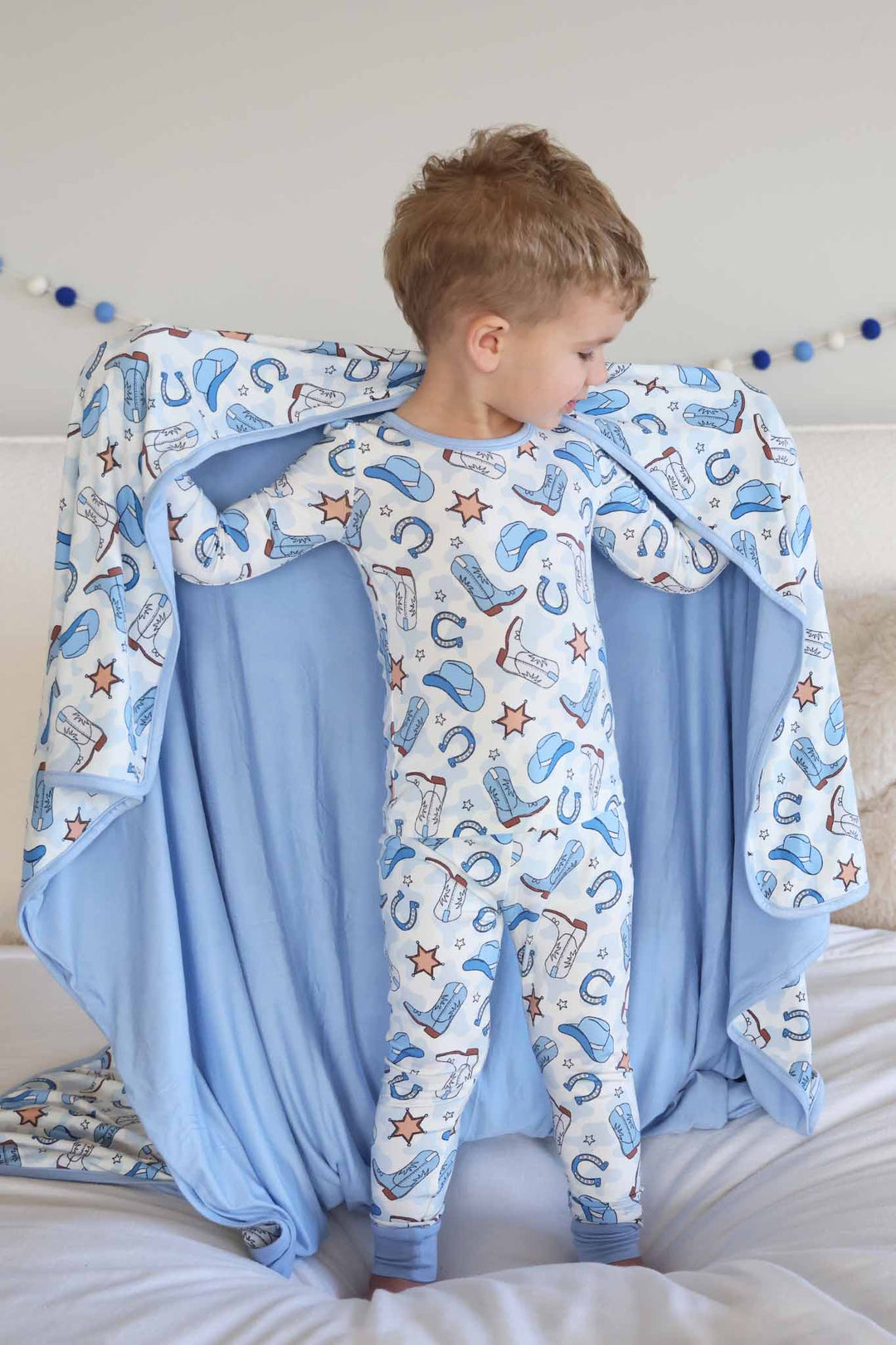 boy standing on bed in blue cowboy themed pajama set holding a matching blanket behind him 