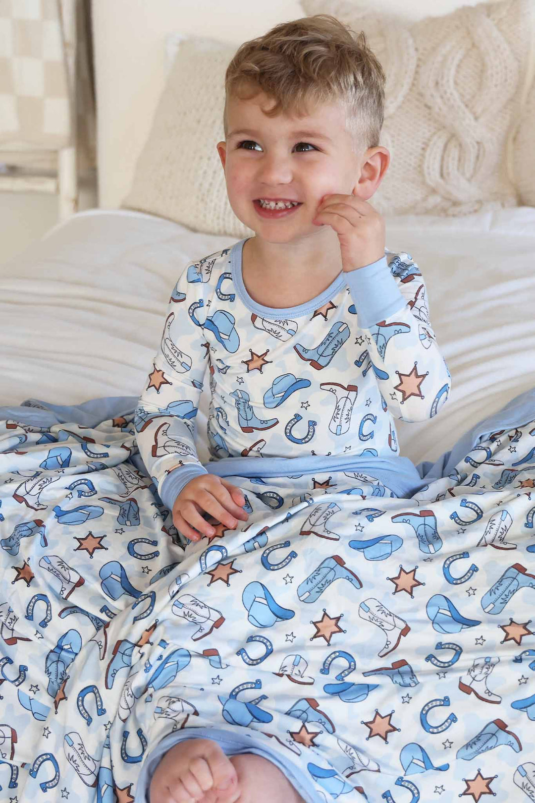 boy sitting on bed wearing a two piece pajama set in a blue rodeo print with a matching blanket across his lap