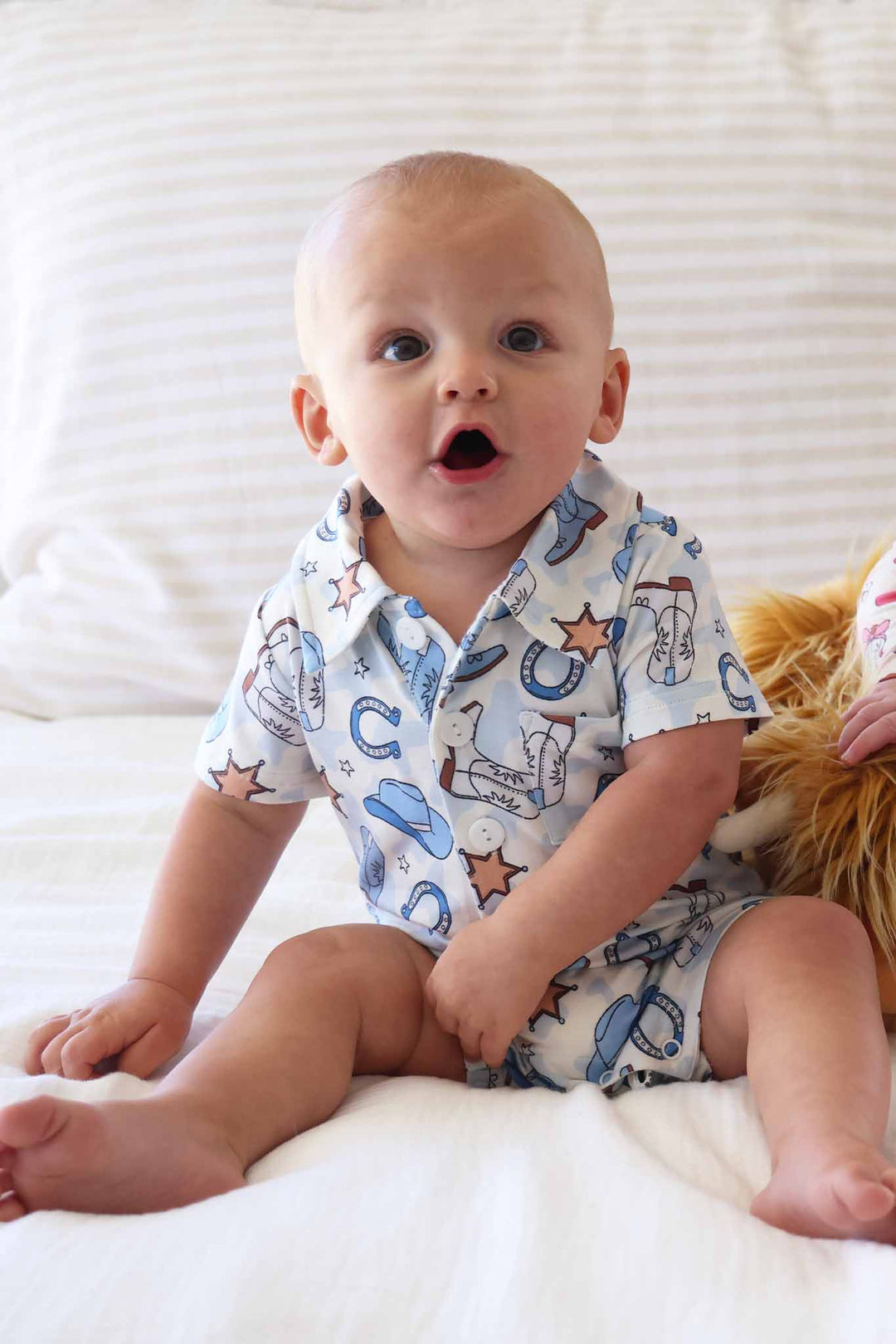 boy sitting on bed in short sleeve collared  button up casual romper in blue rodeo dreams print 