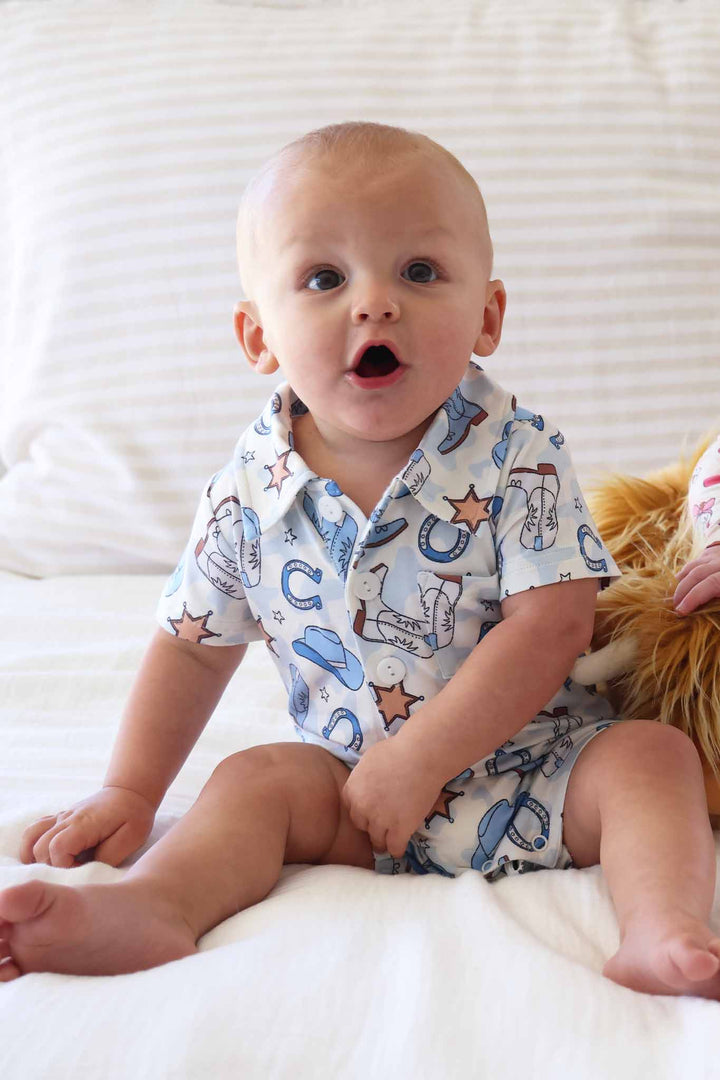boy sitting on bed in short sleeve collared  button up casual romper in blue rodeo dreams print 