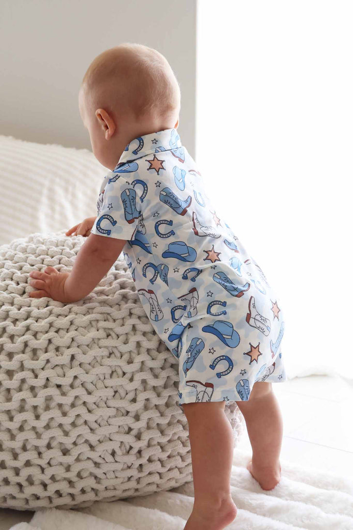standing baby boy in short button romper in blue cowboy print 