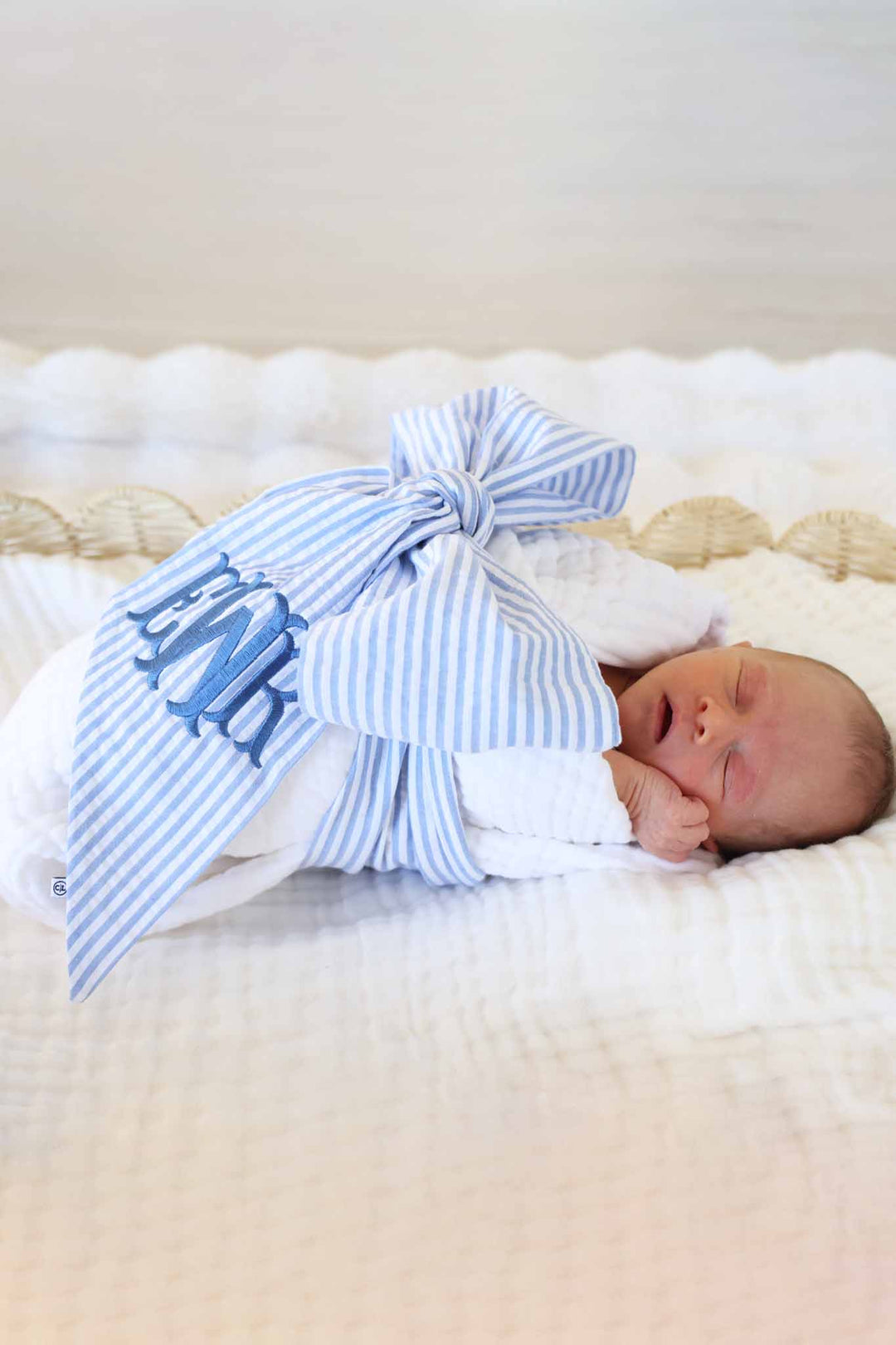 newborn boy wrapped in a swaddle with a blue seersucker bow sash wrapped around him embroidered with monogram