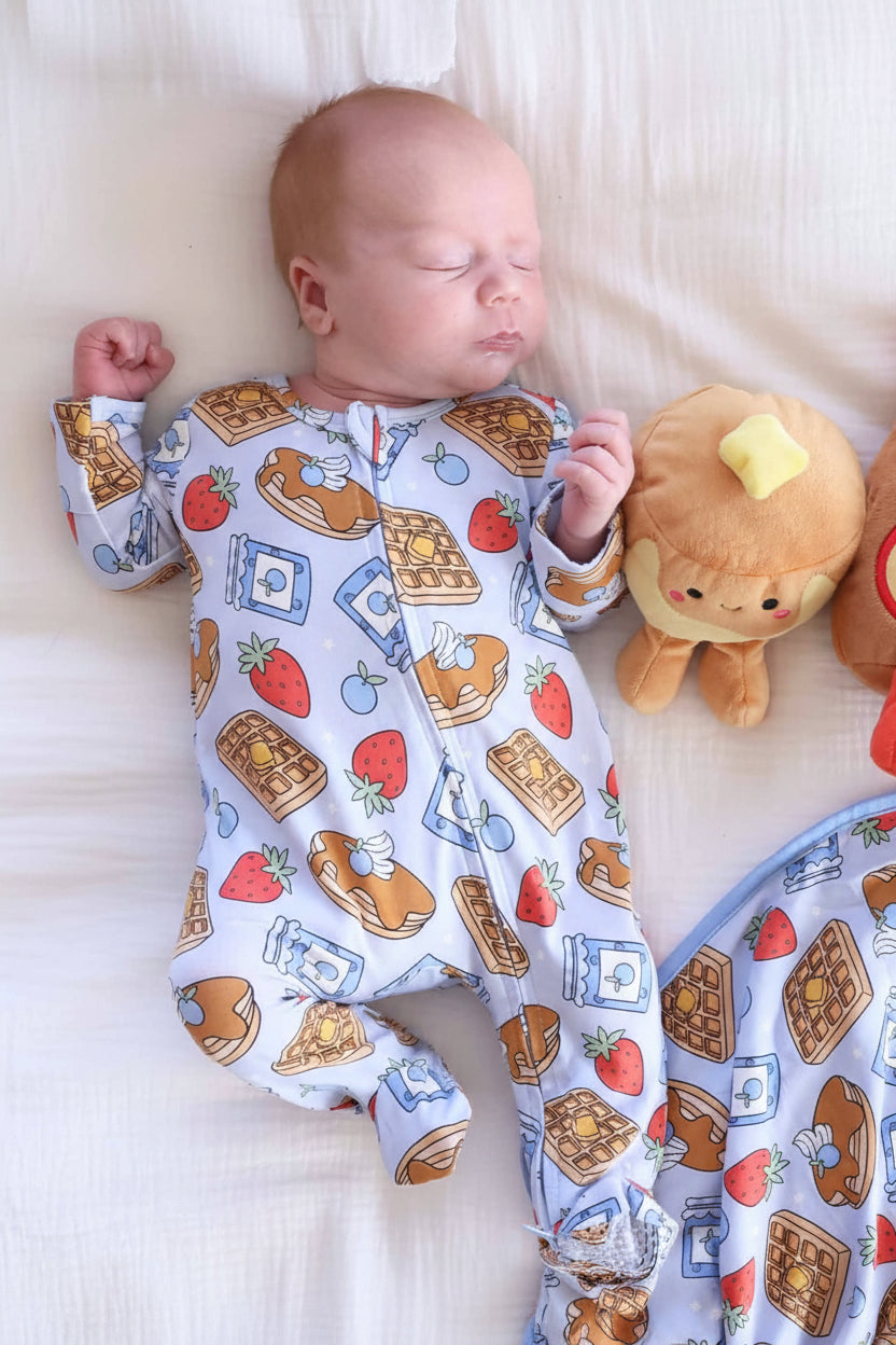 baby boy laying down wearing breakfast themed footie pajama with pancake and syrup stuffed animals