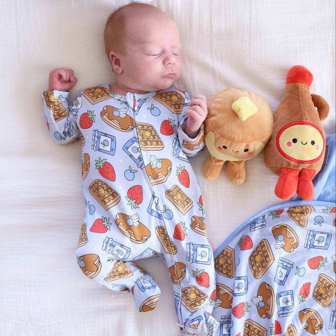 baby boy laying down wearing breakfast themed footie pajama with pancake and syrup stuffed animals