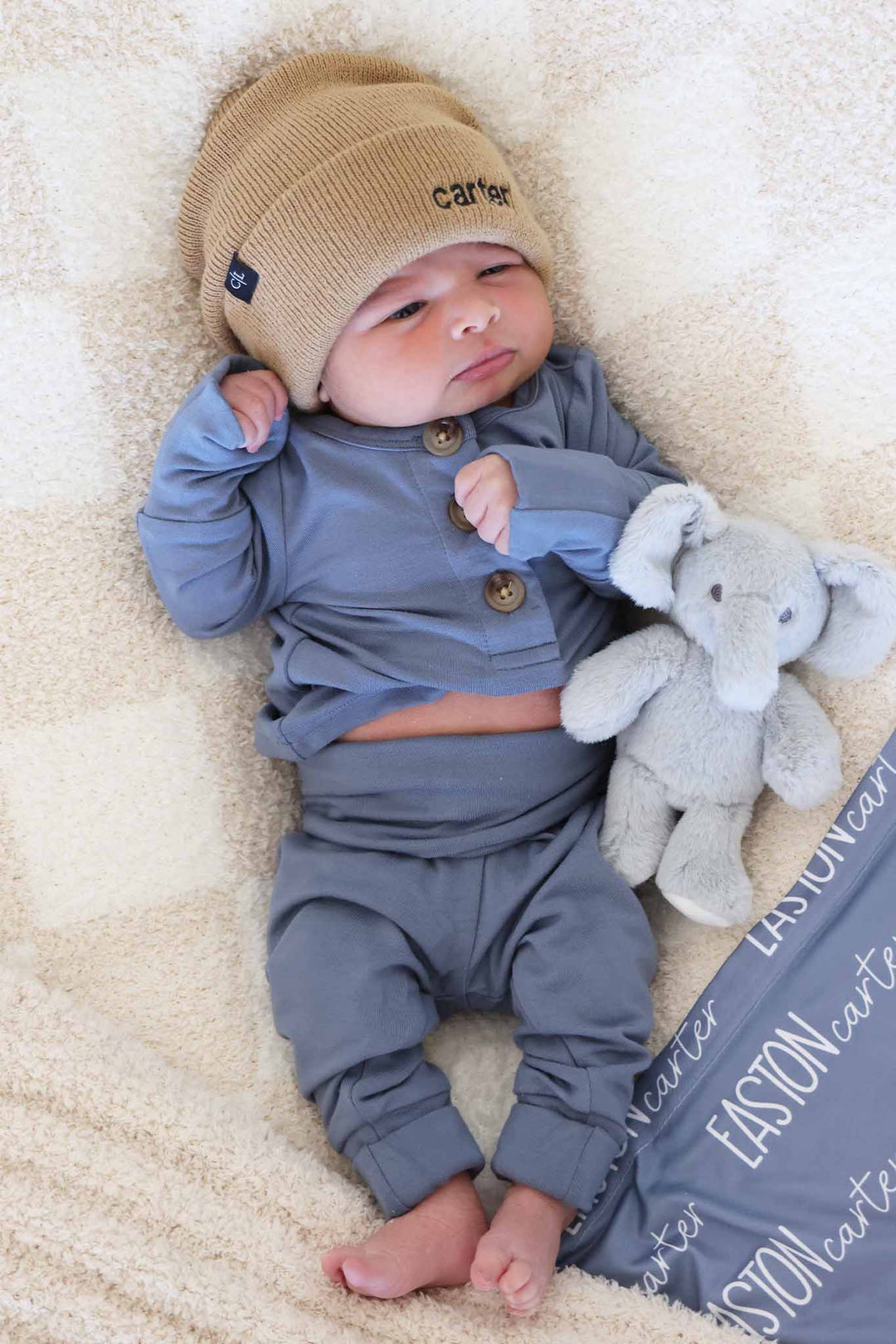 baby boy laying on blanket in blue jean long sleeve and pant two piece set with embroidered beanie and mini elephant stuffed animal 