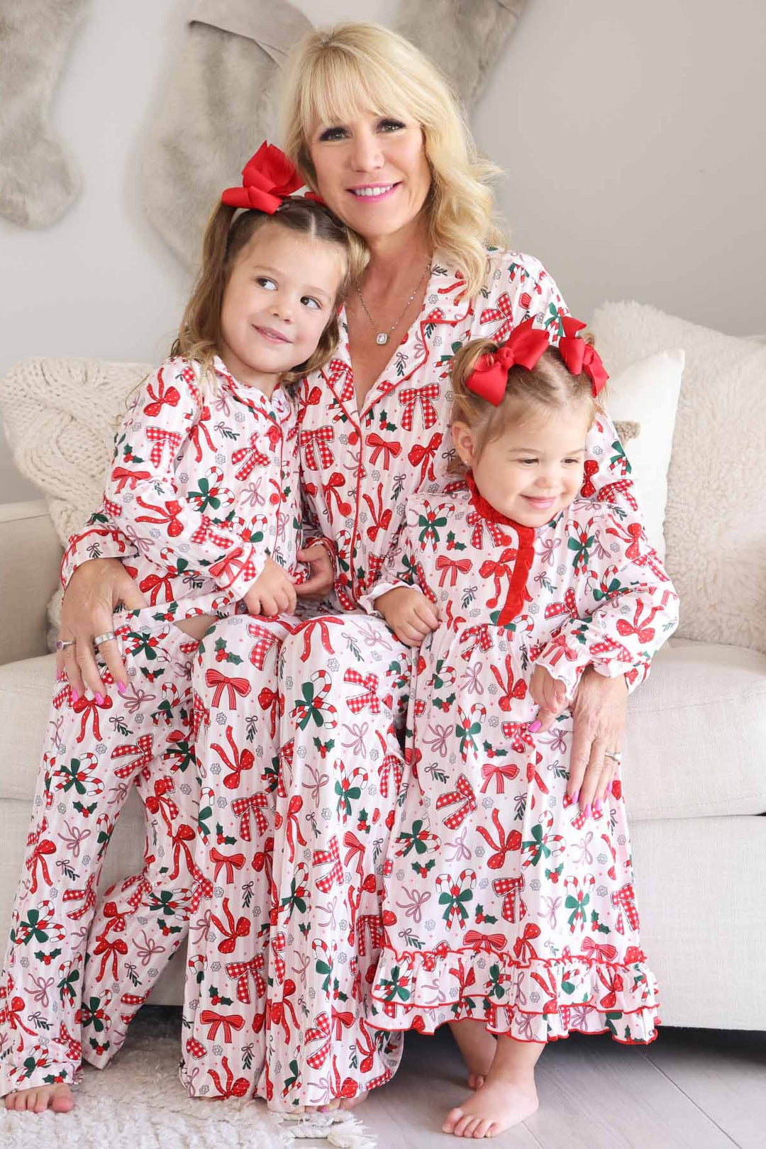 Grandmother and two young girls wearing matching Caden Lane holiday pajamas with festive bow, candy cane, and holly prints while sitting together indoors