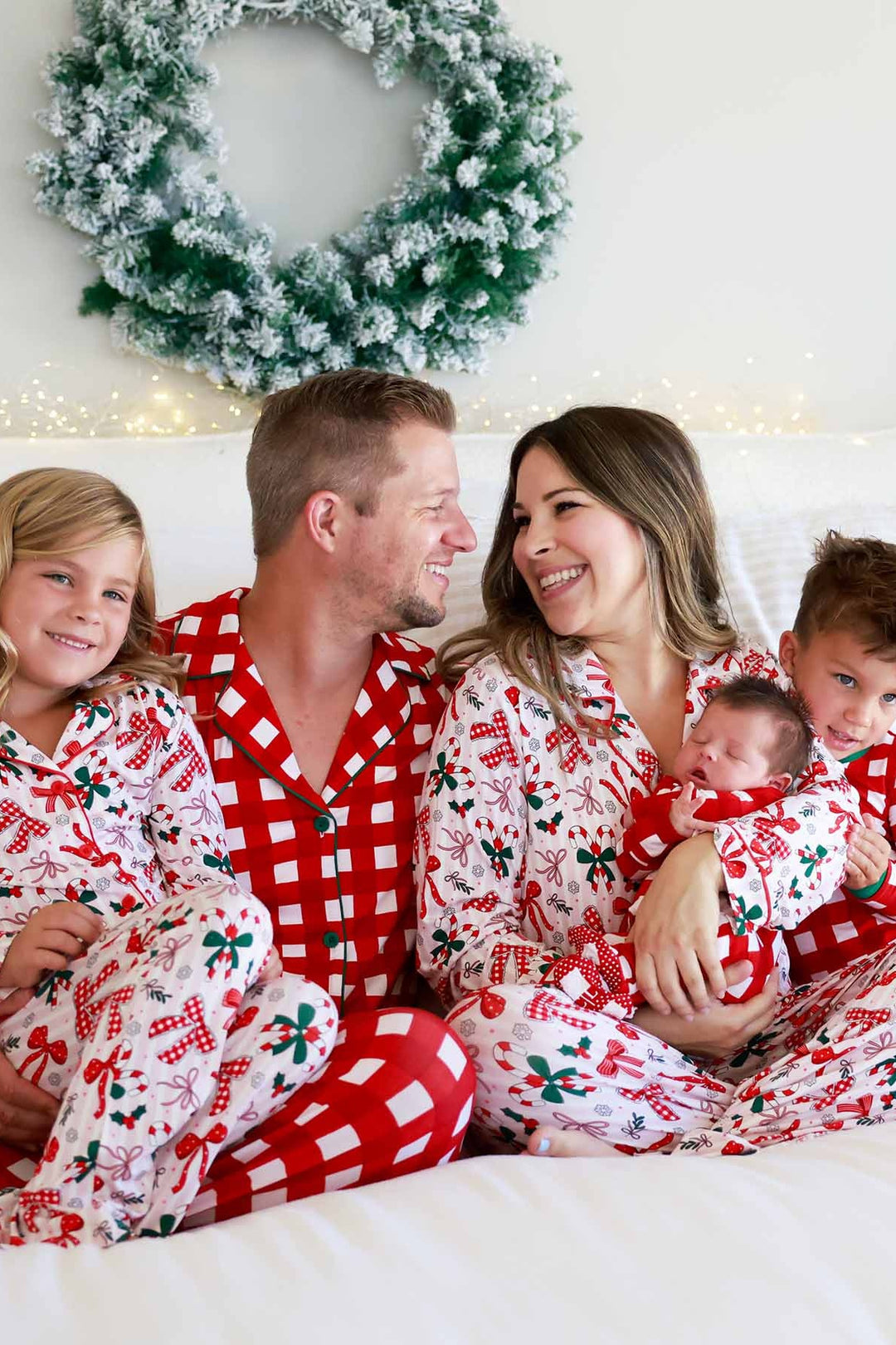 Parents and three children wearing coordinating Christmas Caden Lane pajamas with festive bow prints and red and white checkered patterns, gathered together on a bed beneath a holiday wreath