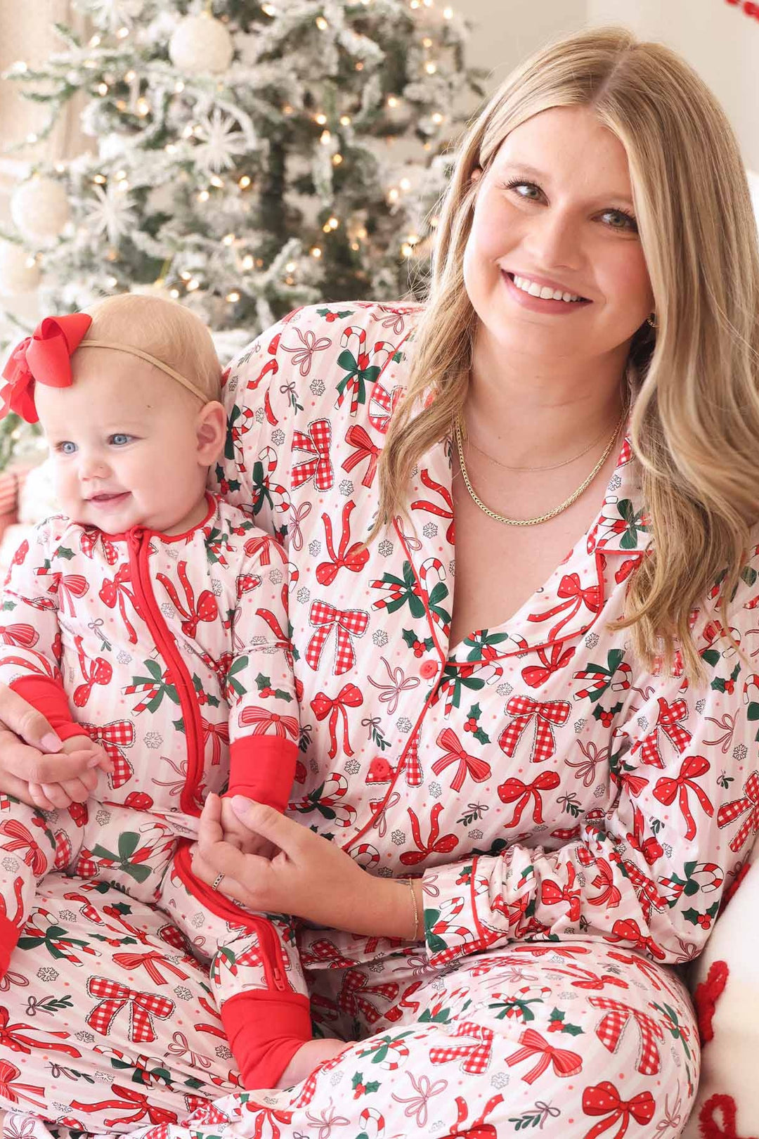 Mother and baby in matching Caden Lane holiday pajamas featuring bows, candy canes, and holly, sitting together in front of a decorated Christmas tree